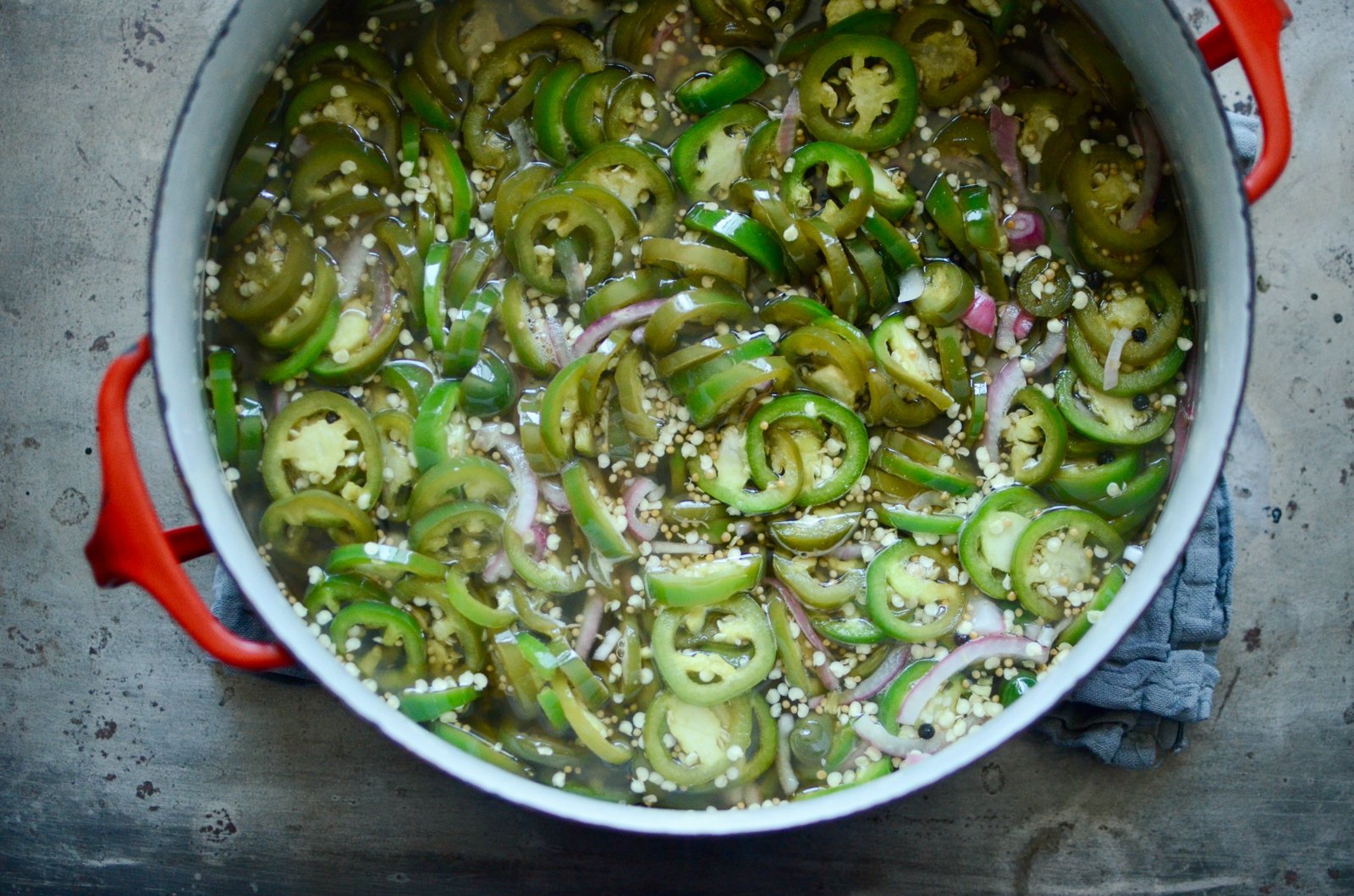 A red enameled pot filled with the cooked pickles. You see the sliced jalapeno rounds interspersed with slivers of red onion. 