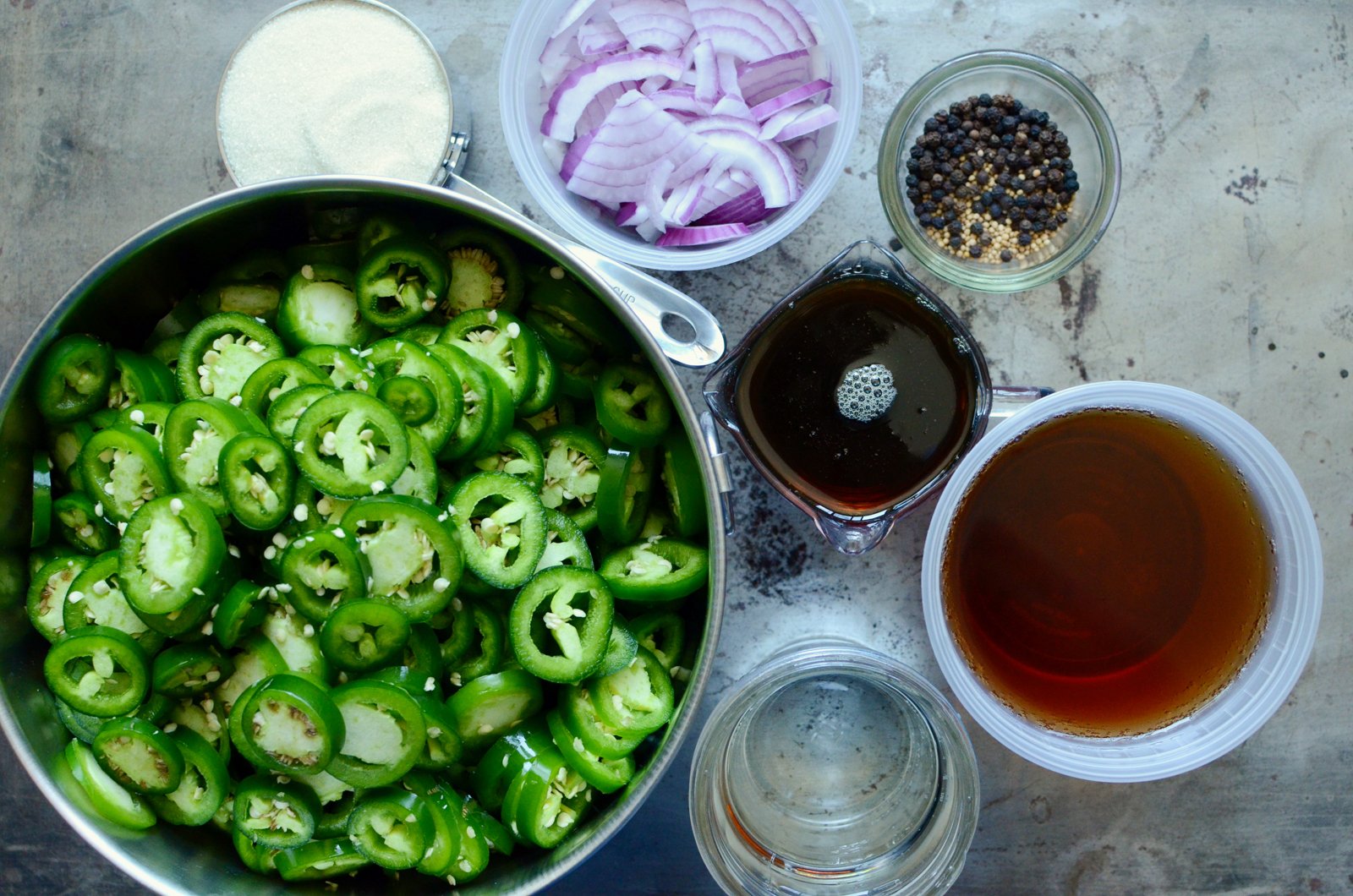 The ingredients for Maple Pickled Jalapenos, resting on a metal sheet pan. Clockwise from the top, there are sliced red onions, a bowl of peppercorns and mustard seed, one cup of maple syrup, three cups apple cider vinegar, water, sliced jalapeno peppers, and one cup of granulated sugar. 